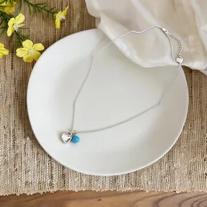 High-angle detail view of a silver heart pendant necklace with turquoise bead on white ceramic