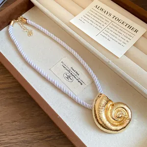 Macro view of a golden metal conch pendant on a braided white silk rope necklace in a box