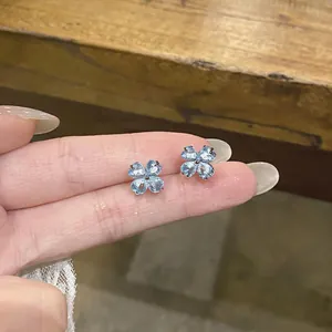 Close-up view of light blue zircon clover stud earrings held between fingers for scale