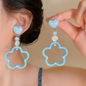 Vista detallada de pendientes de corazón azul y flor de concha usados y sostenidos con la mano