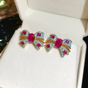 Macro detail of bow-shaped stud earrings with red gemstones and blue pavé stones in a box