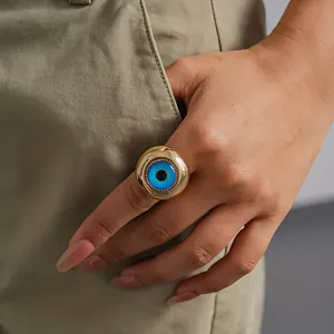 Macro detail of a chunky gold-toned ring with a blue resin evil eye design worn on finger.
