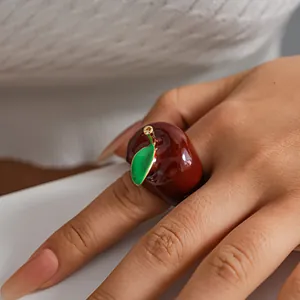 Close-up of a glossy red apple-shaped ring with green leaf detail on a model's finger