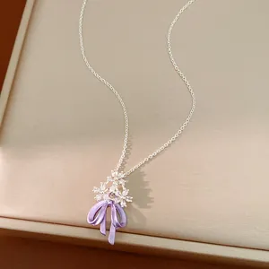 Dettaglio di un ciondolo a fiore in zircone e fiocco in smalto viola su una catena sottile.
