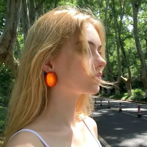 Detail of a large glossy orange oval stud earring worn by a model in a sunlit outdoor setting