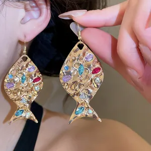 Dettaglio ravvicinato di orecchini a pesce dorati con strass colorati e trama martellata.