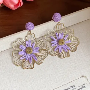 Dettaglio ravvicinato di orecchini a fiore in rafia viola e metallo dorato su scatola crema
