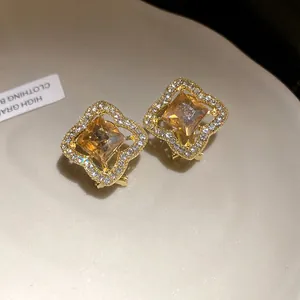 Macro view of gold stud earrings with orange square gem and sparkling stone halo.