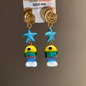 Dettaglio orecchini a tema marino con conchiglia dorata, stella marina blu e pesce colorato