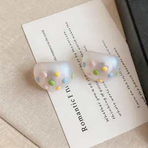 Detalle de pendientes de botón ovalados mate con puntos de colores pastel sobre tarjeta.