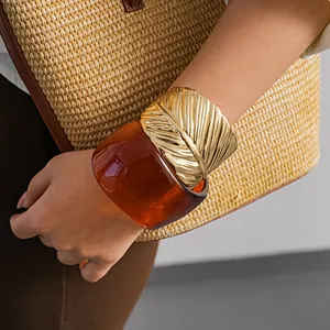 Detailed view of amber resin cuff and gold feather bracelet on wrist with straw bag.