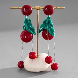 Detailed view of red resin cherry earrings with green leaves and silver tassels on a stand