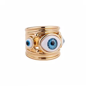 Gold wide band ring featuring blue resin evil eye motifs in a minimalist studio setting