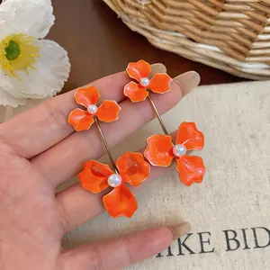 Macro detail of vibrant orange enamel flower drop earrings with pearl centers held by hand.