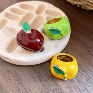 Tres anillos de esmalte de colores con forma de manzana en un soporte crema.