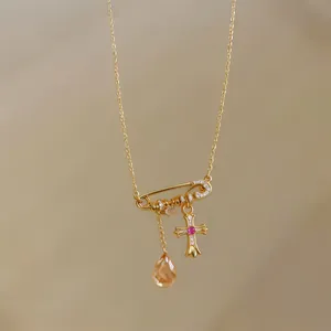 Close-up of a gold safety pin pendant necklace featuring a cross and orange teardrop stone