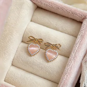 Detailed close-up of pink cat-eye heart stud earrings featuring a gold bow and sparkling rhinestones