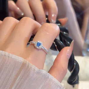 Close-up of a silver ring with a square blue gemstone and crystal halo on a finger