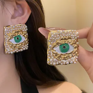 Close-up of vintage square stud earrings with pearl borders and green enamel eye detail.