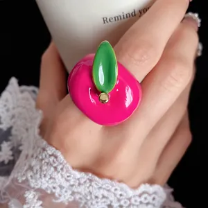 Close-up of a vibrant pink strawberry enamel ring with a green leaf on a finger.