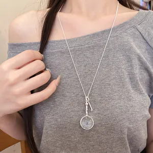 Close-up of a long silver necklace with a vintage circular magnifying glass key pendant on a model.