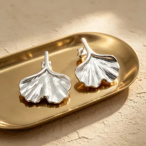 Detailed close-up of silver ginkgo leaf earrings resting on a golden tray in natural sunlight