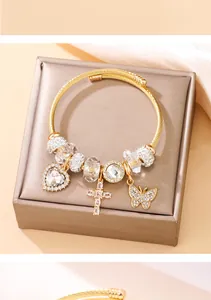 Vue détaillée d'un bracelet à breloques en acier doré avec cœur, croix et papillon sur boîte