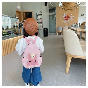 Sac à dos rose pour enfants avec un ours en peluche 3D dans un décor de café lumineux.