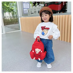 Sac à dos rouge pour enfant avec décoration ours en peluche tenu par une petite fille.