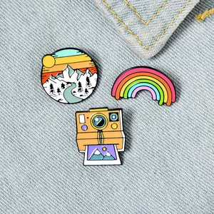 Close-up of camera, rainbow, and sunset enamel pins pinned to a light denim jacket