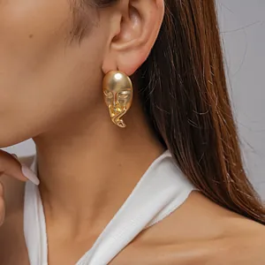 Close-up of matte gold mask earring showing the surrealist portrait detail on model