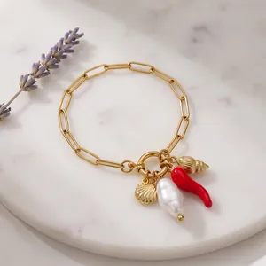 Gold paperclip chain bracelet with shell, pearl, and red chili charms on marble base.