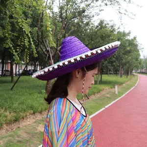 Large purple straw Mexican sombrero with white pom-poms worn by a person in a park.