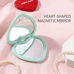 Portable mint green heart-shaped magnetic flip makeup mirror on soft pink fabric.