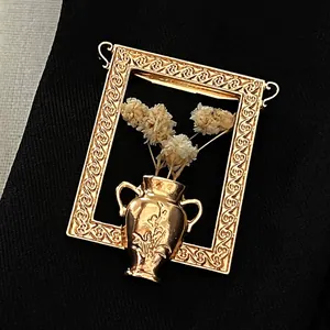Nahaufnahme einer goldfarbenen Rahmenbrosche mit Miniaturvase und weißen Trockenblumen.