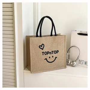 Jute tote bag with TOPnTOP smiley print on white tabletop with silver headphones.