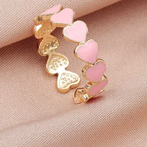 Detalle de anillo ajustable con corazones de esmalte rosa y acabado dorado sobre tela rosa.
