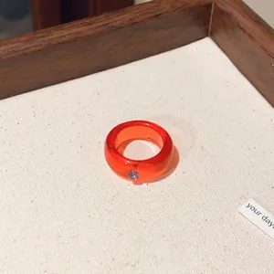 Detailansicht eines orangefarbenen Harzrings mit kleinem lila Stein auf beigem Hintergrund.