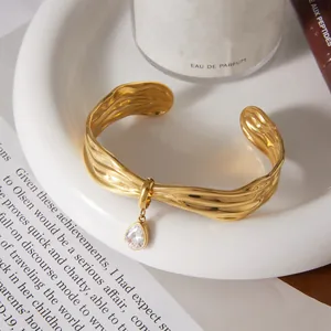 Close-up detail of a wavy gold cuff bracelet with a teardrop-shaped zircon charm.