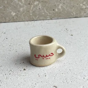 Macro view of a miniature beige ceramic mug charm with red Habibi Arabic script engraving