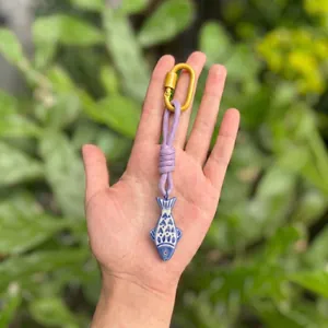 Detailed view of a blue ceramic fish charm keychain with purple rope and a gold carabiner clip.