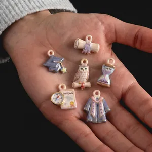 Hand holding six miniature graduation-themed ceramic charms with gold accents