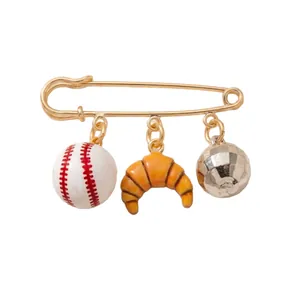 Gold safety pin brooch with baseball, croissant, and disco ball charms on white background