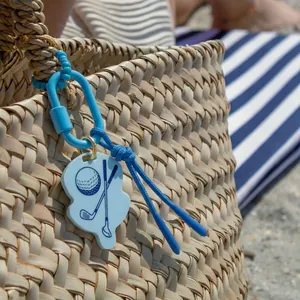 Close-up of a blue golf-themed charm attached to a woven straw bag on the beach