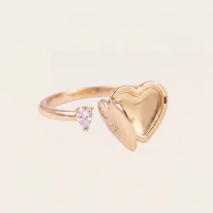 Detalle de anillo abierto con relicario de corazón dorado y piedra rosa en gota