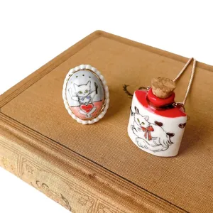 Healing cat-themed ceramic jewelry set with painted bottle necklace and ring on vintage book.