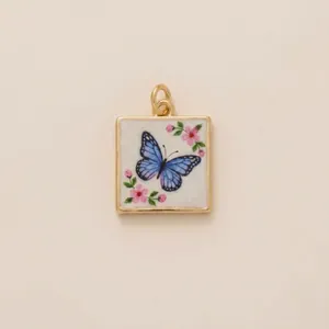 Macro detail of a square gold pendant featuring a blue butterfly and pink flowers.