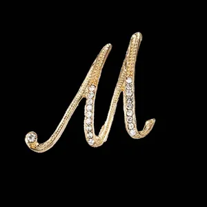 Detalle de un broche dorado en forma de letra M cursiva con incrustaciones de strass