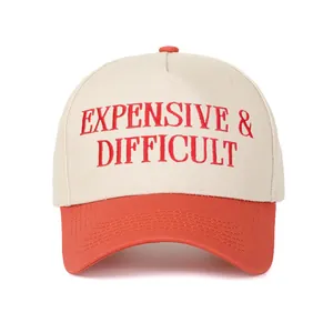 Κοντινό πλάνο κόκκινου κεντήματος 'Expensive & Difficult' στο μπεζ-πορτοκαλί καπέλο