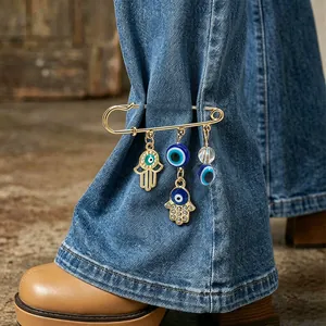 Close-up detail of a gold Hamsa hand and Evil Eye charm pin on denim jeans leg.